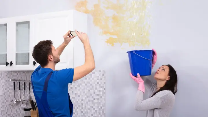 Two people looking at a water stain on a wall with a bucket catching drips